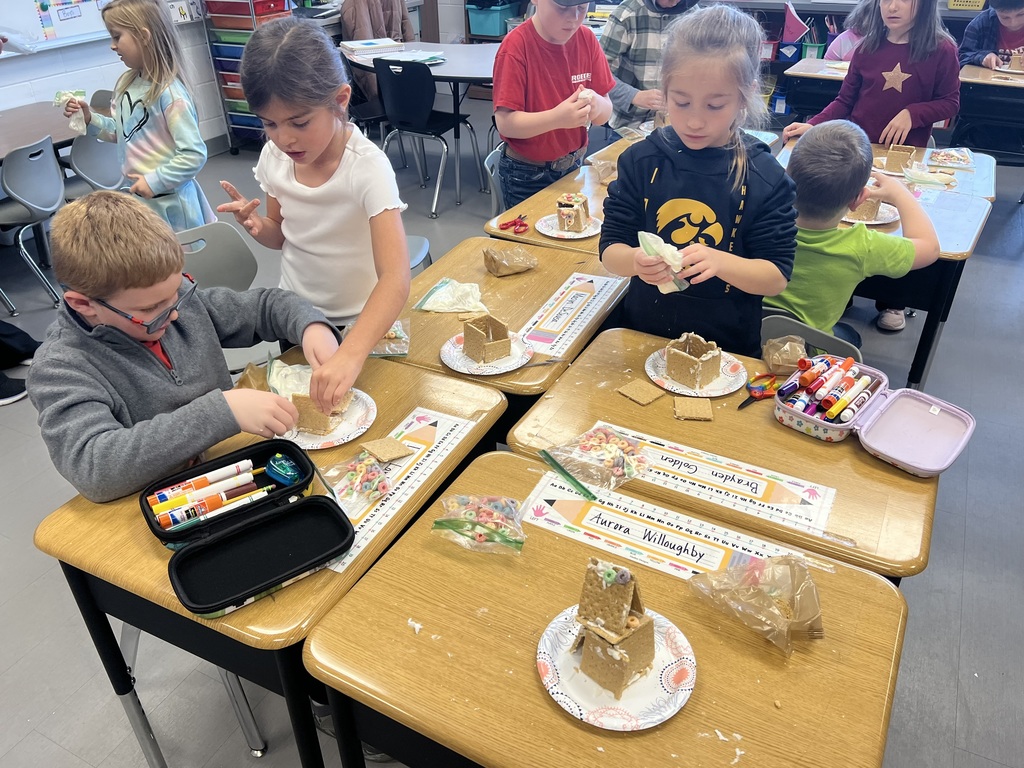 Students gather at their desks constructing graham cracker houses using icing and cereal, with school supplies nearby.
