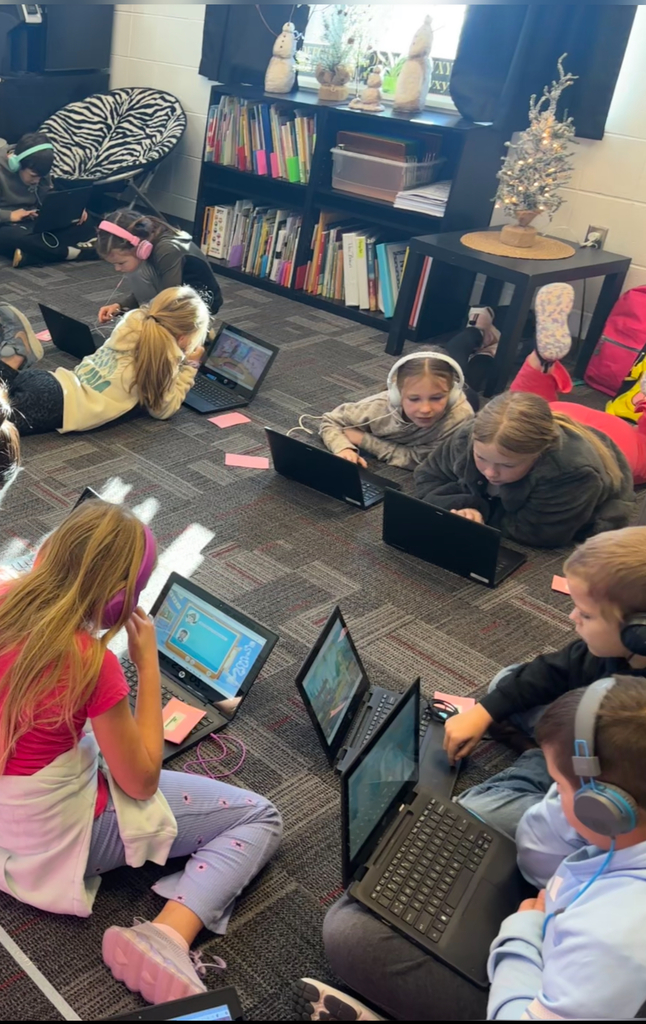 A group of students wearing headphones work on laptops while seated and lying on the classroom floor during a tech activity.