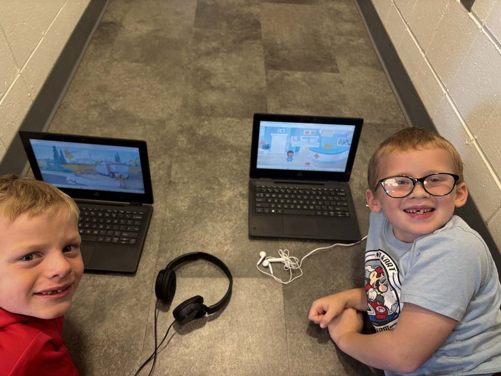 Two young boys smile while sitting on the floor with laptops and headphones, each playing educational games.