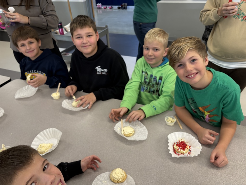 Smiling boys sit at a table with cookies, frosting, and paper liners, enjoying a cookie decorating activity.