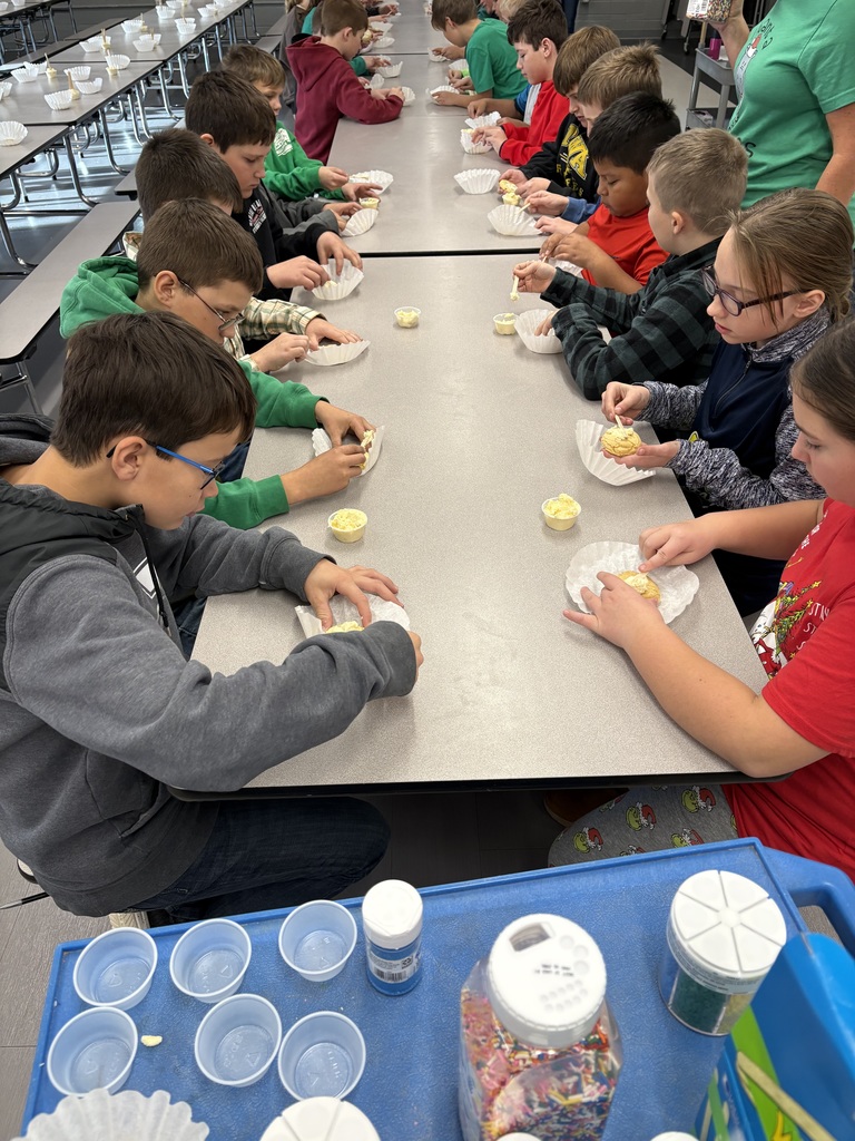 Students sit at cafeteria tables decorating cookies with frosting, using paper liners and plastic utensils.