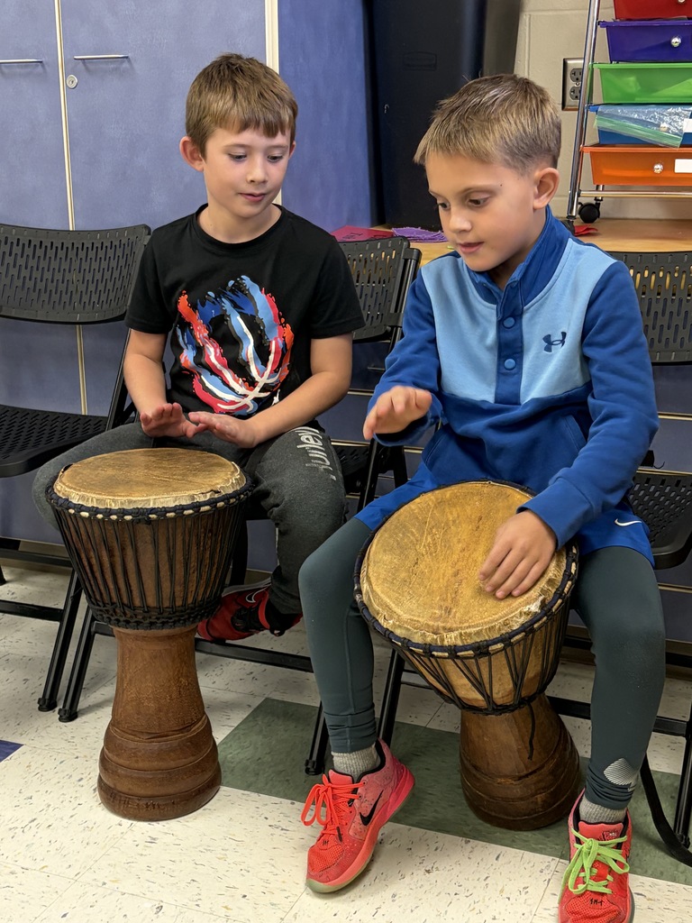 Two boys sit side by side, focused on playing djembe drums together during a classroom music lesson.