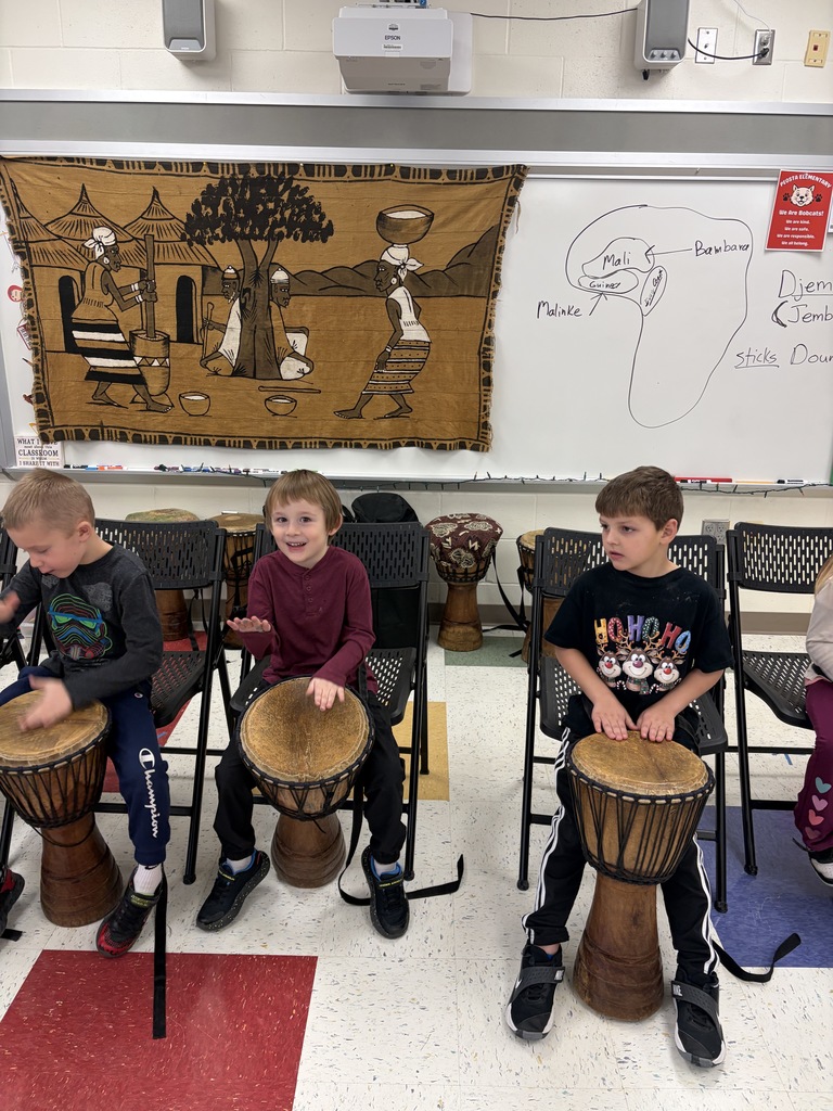 Three young boys sit in a classroom, each playing a djembe drum, with a cultural tapestry and map of Africa in the background.