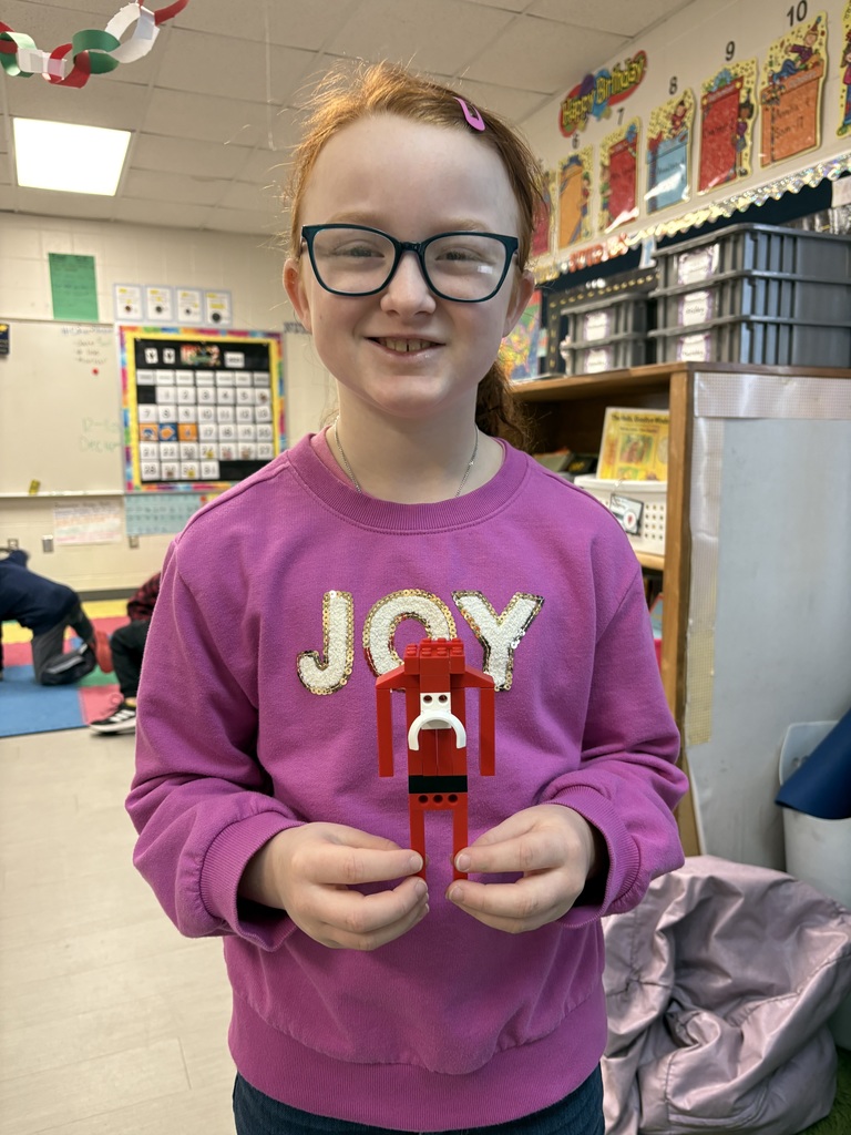 A smiling girl in a purple “JOY” sweatshirt holds a red LEGO Santa figure in a colorful classroom setting.