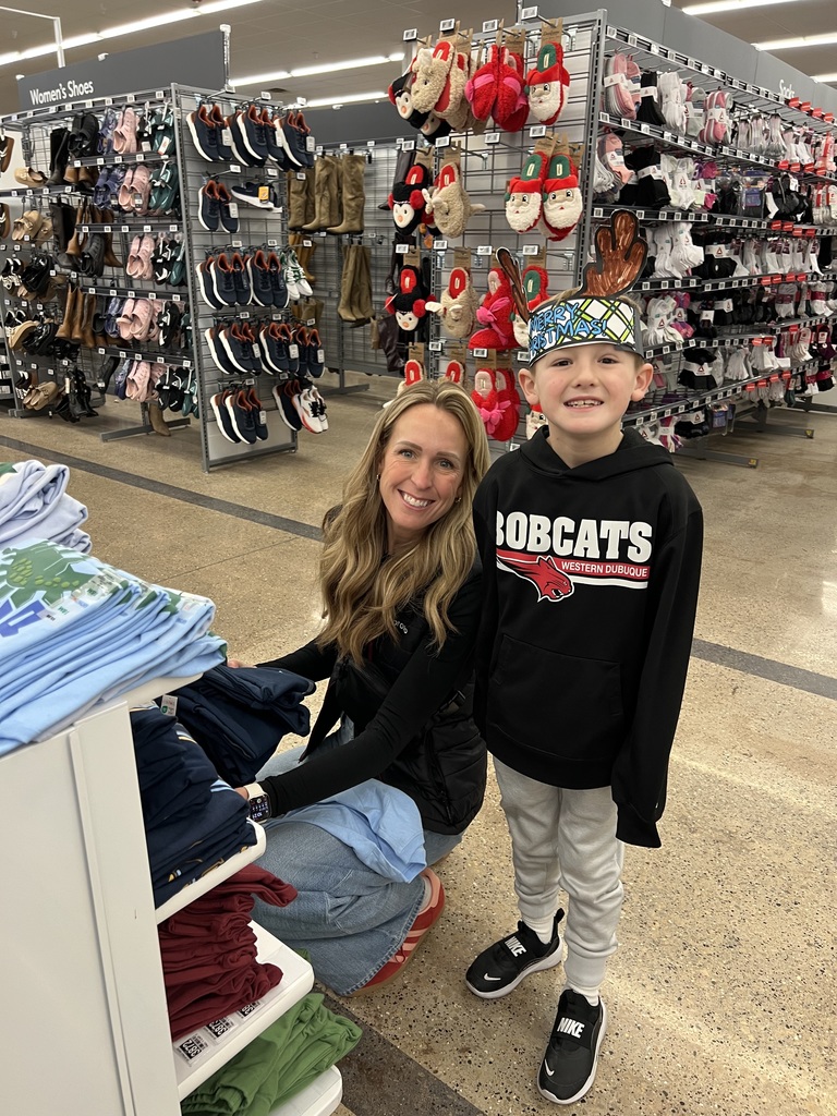 A woman kneels beside a young boy wearing a reindeer hat in a clothing section of a store, both smiling next to a display of folded shirts and racks of slippers.