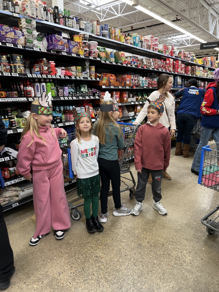A group of five children wearing holiday reindeer hats stand in a grocery aisle near carts and shelves filled with snacks and packaged foods during a festive shopping trip.