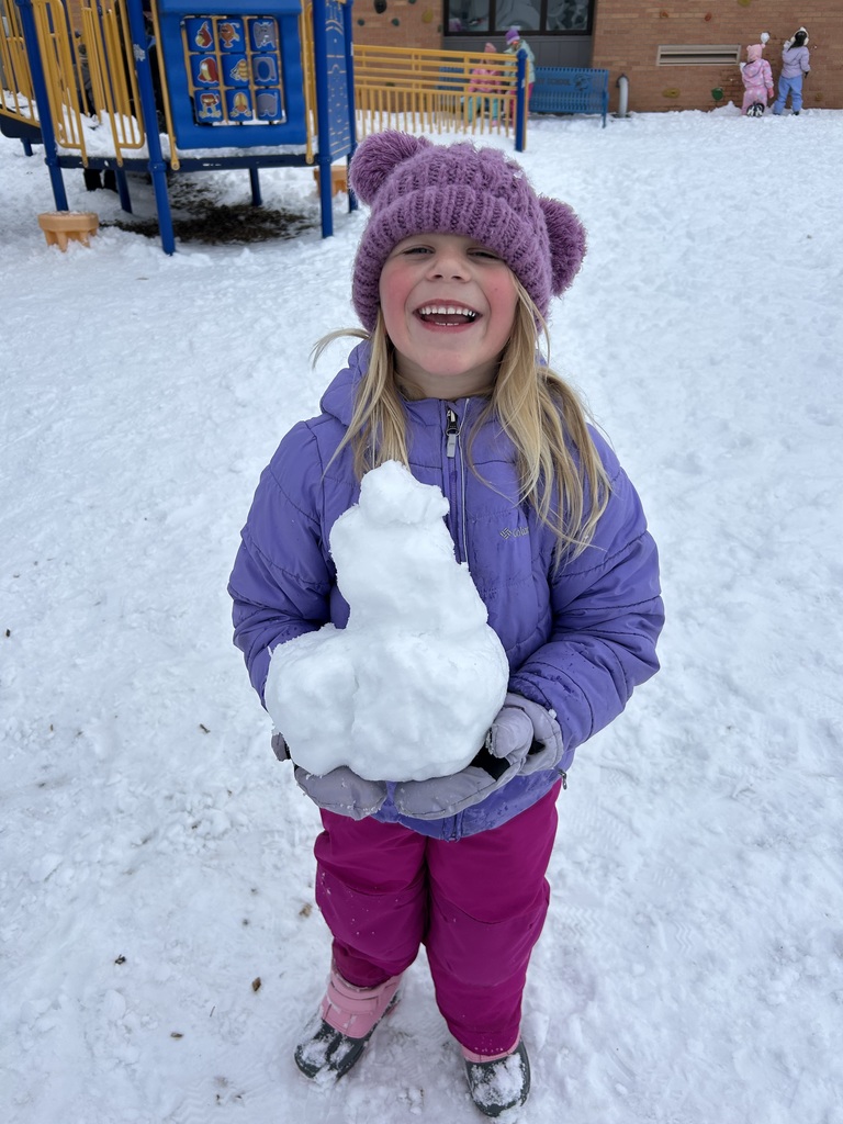 A smiling girl in a purple hat and jacket proudly holds a small snowman in the school playground, surrounded by snow and colorful playground equipment.