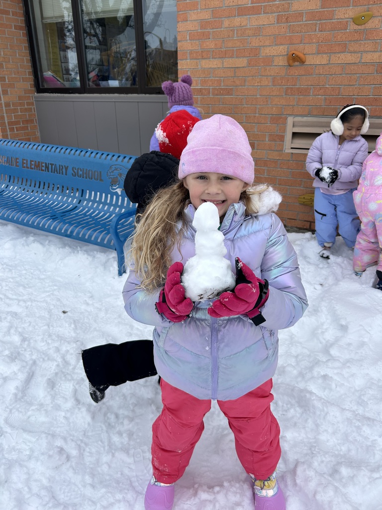 A young girl in a pink hat and gloves holds a small snowman she built, smiling outside Cascade Elementary School as other children play in the snow nearby.