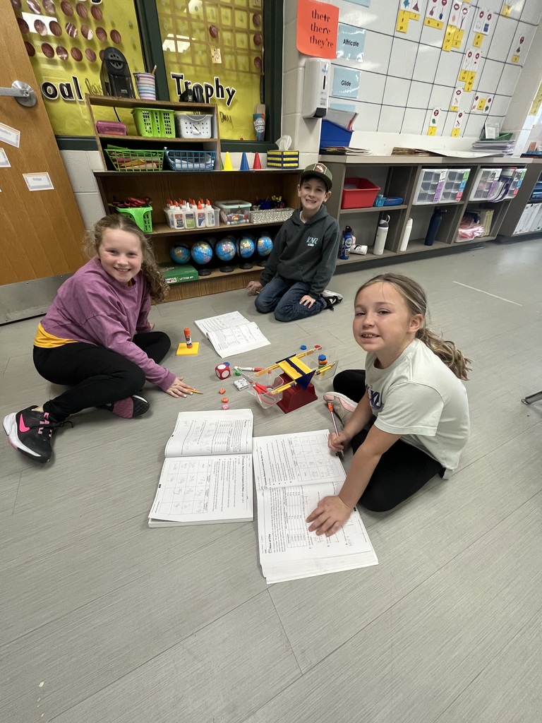 Three smiling students sit on the classroom floor working on activity books and using math manipulatives, surrounded by educational supplies and colorful decorations.