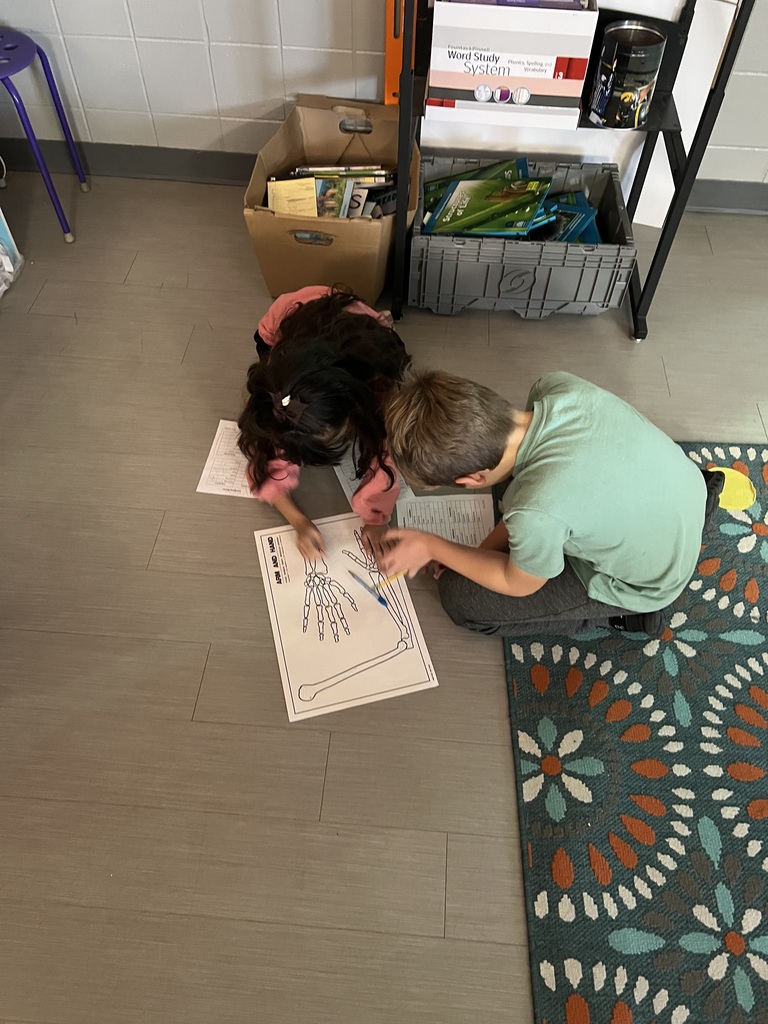 Two students sit on the classroom floor working together on a worksheet featuring a diagram of a human hand skeleton, surrounded by school supplies and a colorful floral rug.