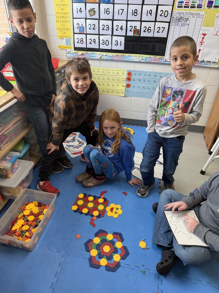 A group of students sit and kneel on a blue mat, proudly displaying intricate designs they created using colorful pattern blocks in the classroom.