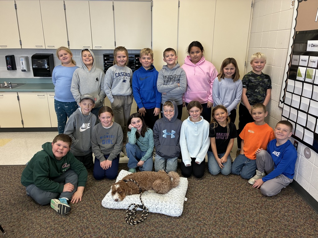 A group of smiling elementary school students pose together in a classroom, with a brown and white therapy dog resting on a soft bed in front of them.