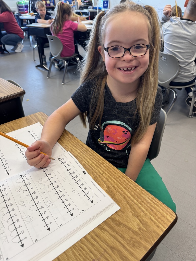 A young girl with glasses and pigtails smiles while sitting at a classroom desk, working on a number line math worksheet with a pencil.
