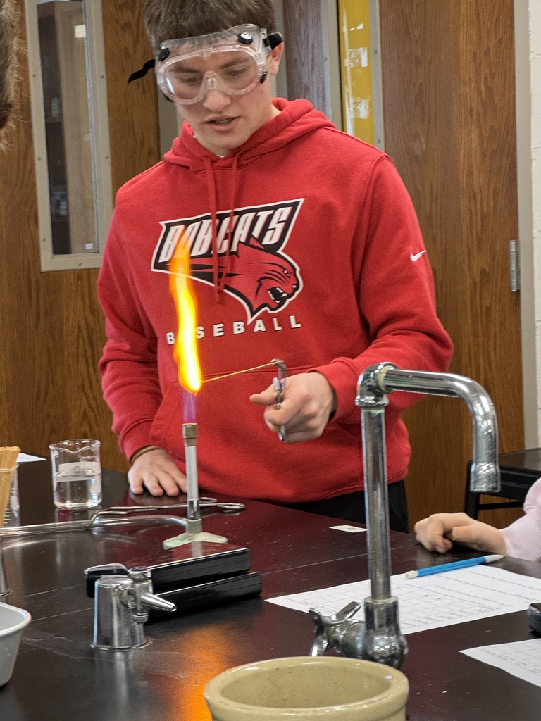 A student in a red “Bobcats Baseball” hoodie and goggles holds a splint with a tall orange flame over a Bunsen burner during a chemistry experiment in class.