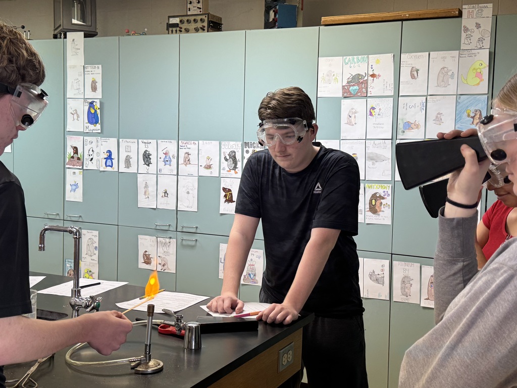A group of students in a science lab perform a flame test. A Bunsen burner with an orange flame is on the table.