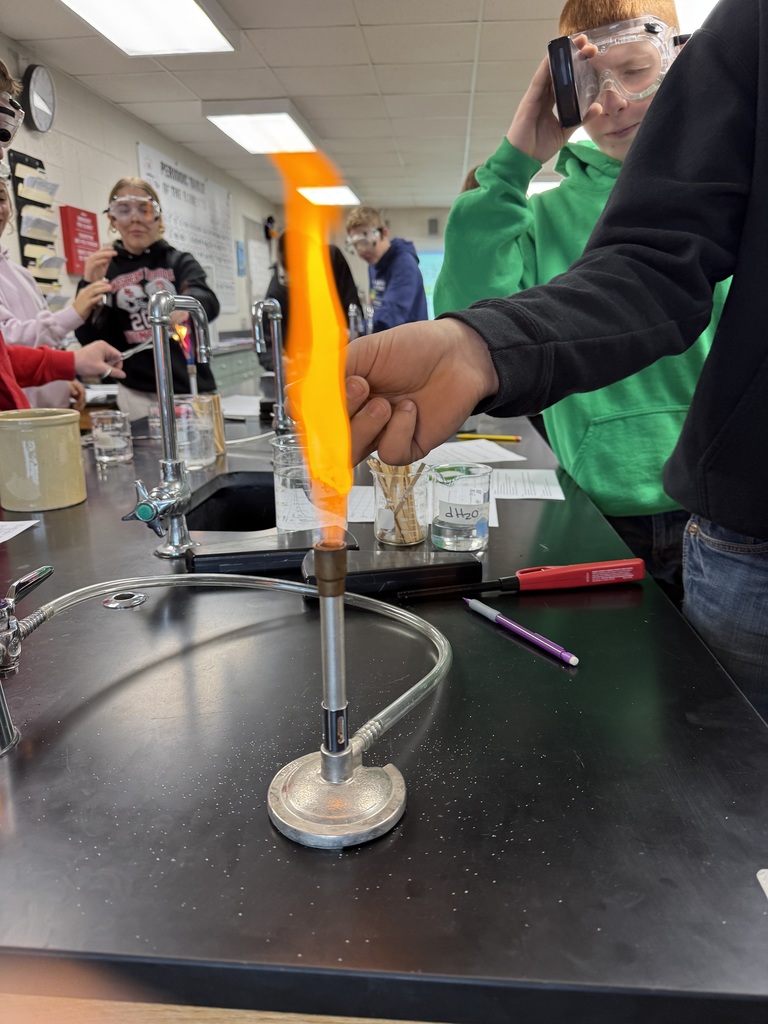 A close-up view of a student’s hand holding a splint in a bright orange flame during a science lab flame test.