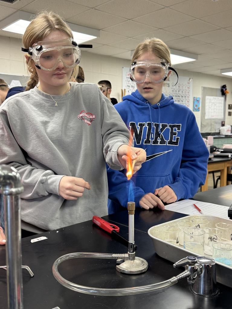 Two students wearing goggles observe a flame test in a science lab. One student holds a splint with an orange flame over a Bunsen burner while the other watches closely.