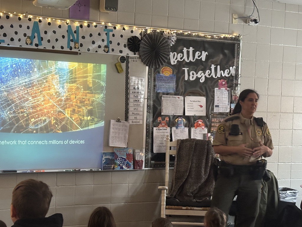 The officer speaks to a classroom of children, standing near a screen showing internet-related graphics and a "Better Together" bulletin board.