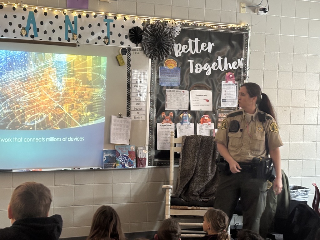 A uniformed officer stands beside a classroom projector screen displaying digital network imagery, speaking to seated students.