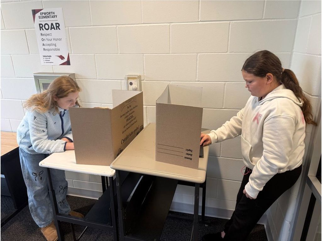 Two students focus on their laptops at separate desks, each using cardboard dividers for privacy near a wall sign reading “Epworth Elementary ROAR.”
