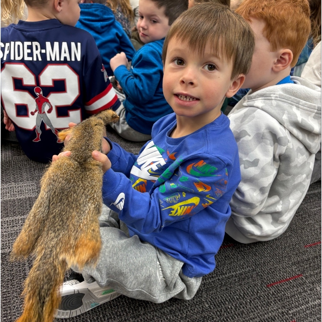 A boy holds a preserved animal pelt, possibly a squirrel, while sitting among classmates and smiling at the camera.
