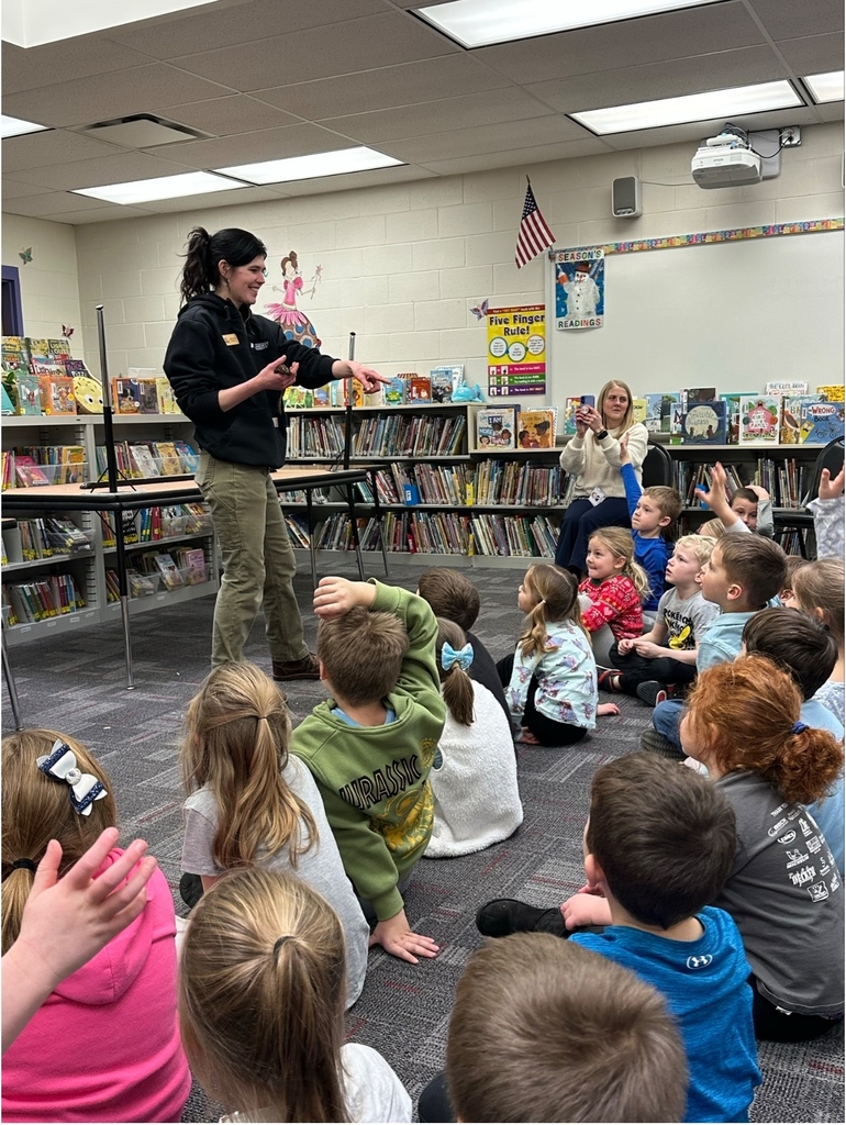 A woman smiles while holding a small lizard or reptile on her hand during a classroom demonstration.