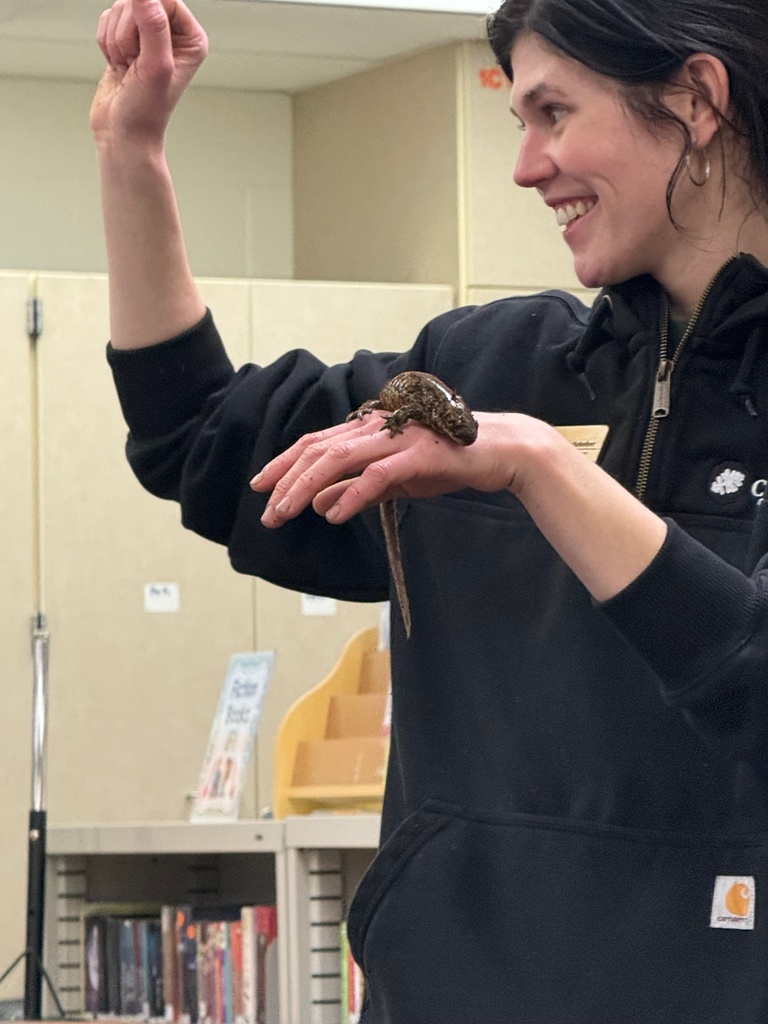 A woman smiles while holding a small lizard or reptile on her hand during a classroom demonstration.