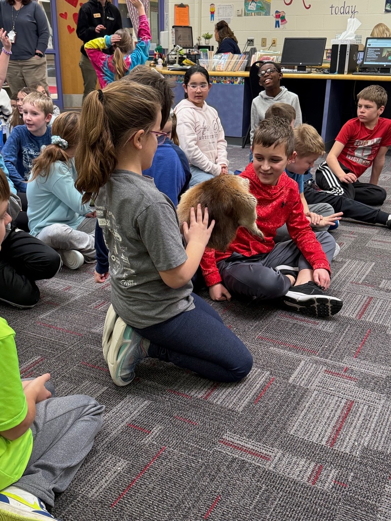 A girl shows an animal pelt to a seated classmate during an educational session in a library.