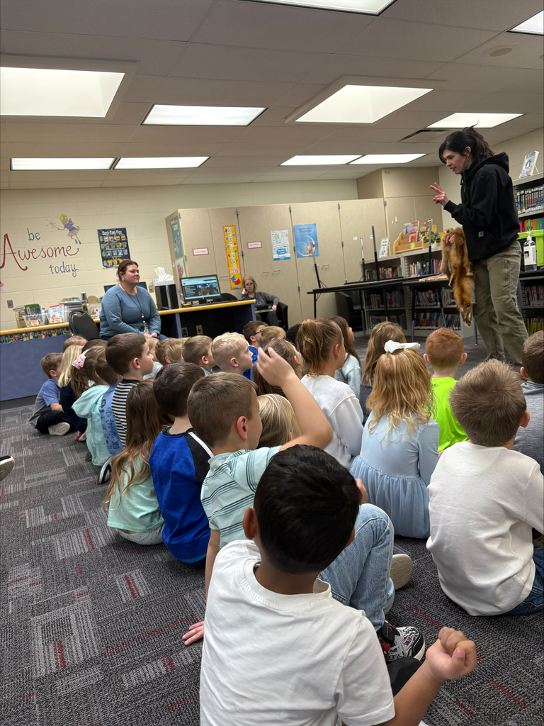 A woman stands at the front of a library classroom holding animal pelts as students sit on the floor watching and raising hands.