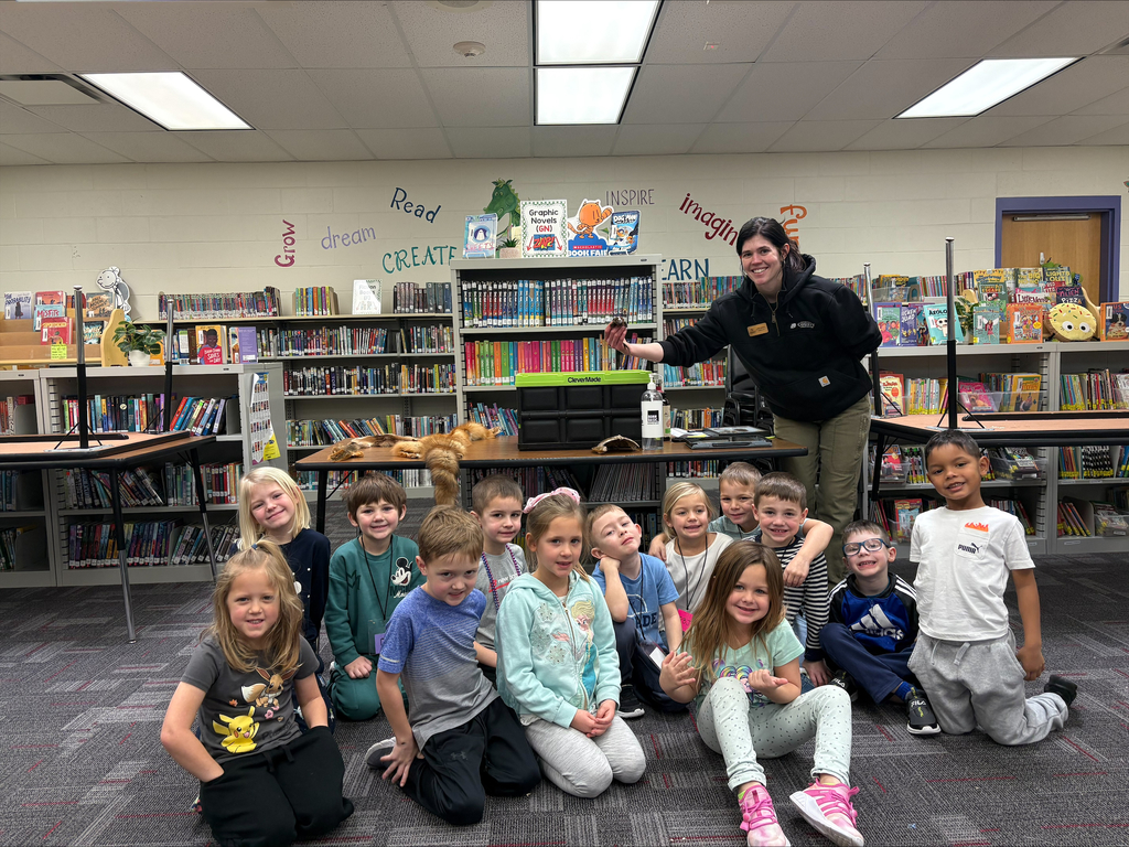 A group of children poses smiling with an animal educator in a school library, with animal pelts on the table behind them.