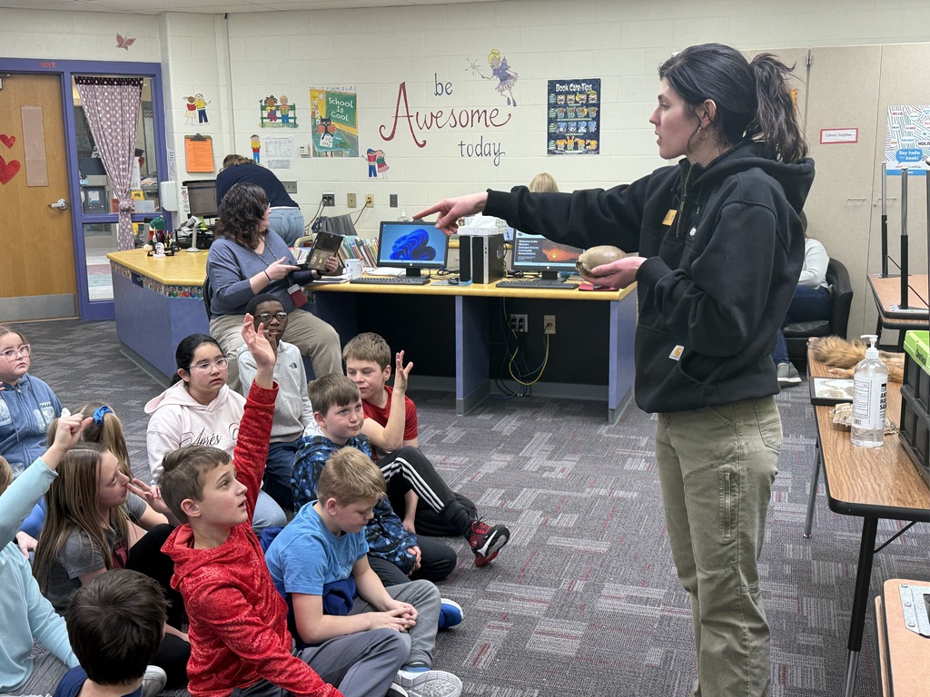 A woman holds a small armadillo shell while presenting to a group of students, some raising hands, in a school library.