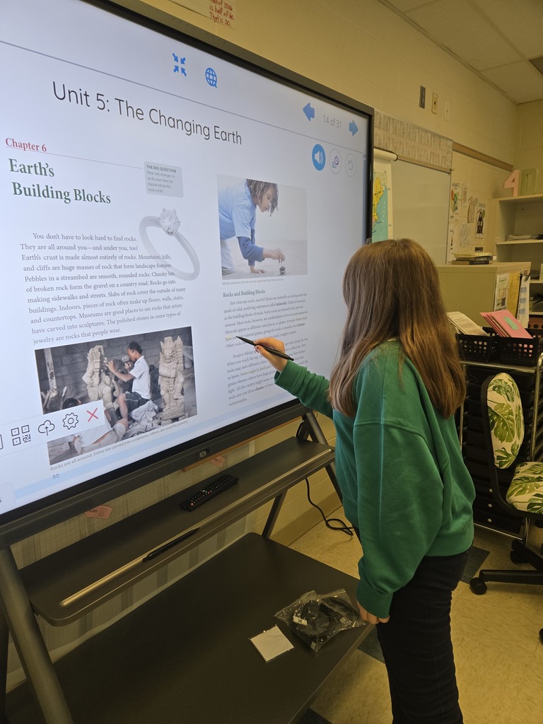 A girl in a green sweatshirt interacts with a large digital screen during a geology-themed reading lesson.