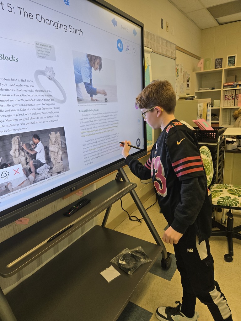A boy in a sports jersey writes on a touchscreen display during a classroom lesson about Earth's rock types.