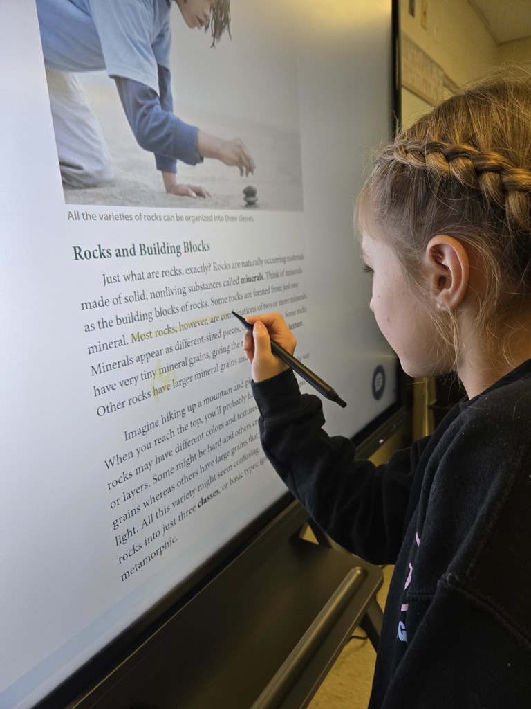 A student with braided hair traces words on a digital display during a lesson on rocks and building blocks of Earth.