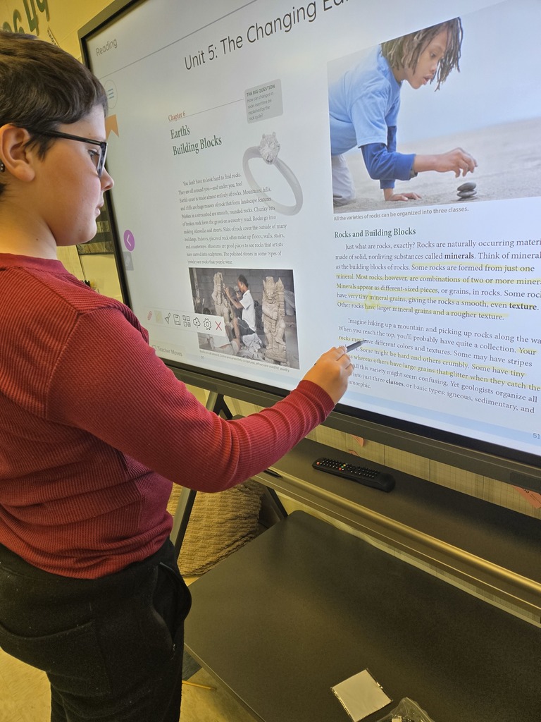 A student in a red shirt interacts with a digital lesson about Earth’s rocks and minerals on a large touchscreen display.