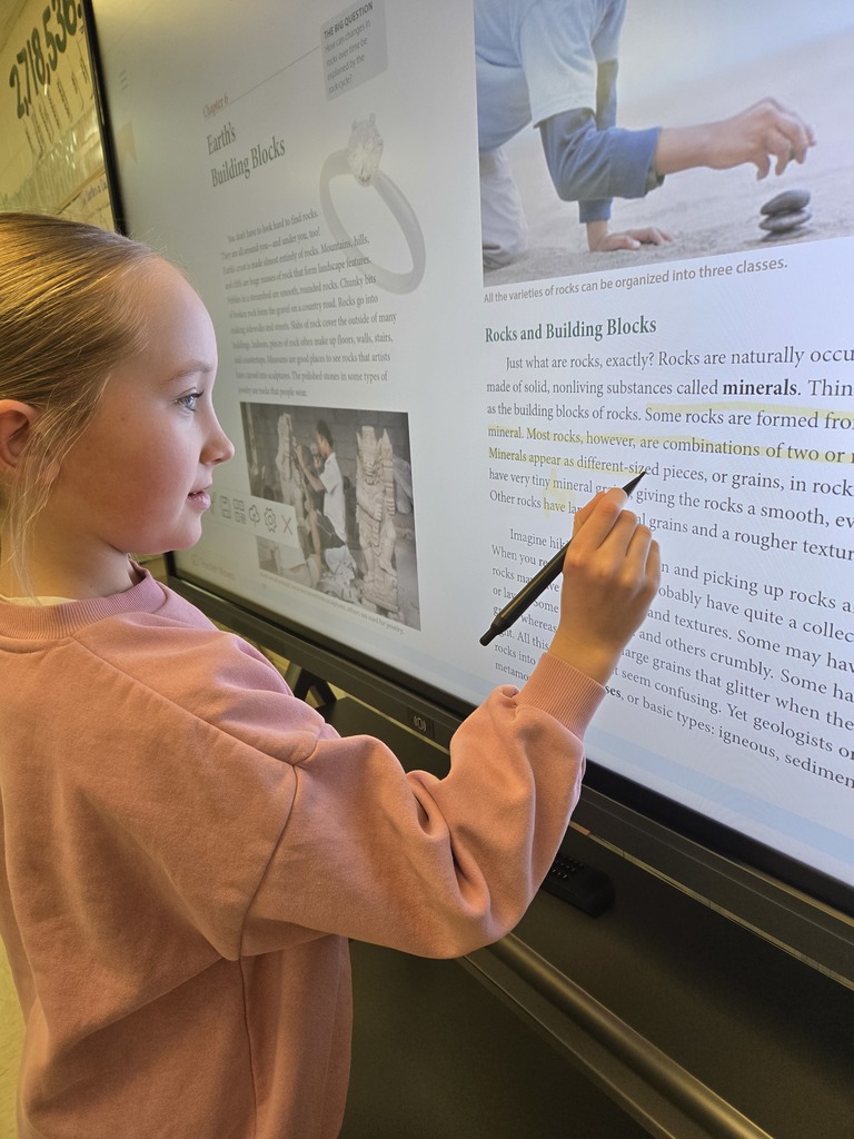 A girl uses a stylus to highlight text on a digital screen showing a science lesson about rocks and minerals.