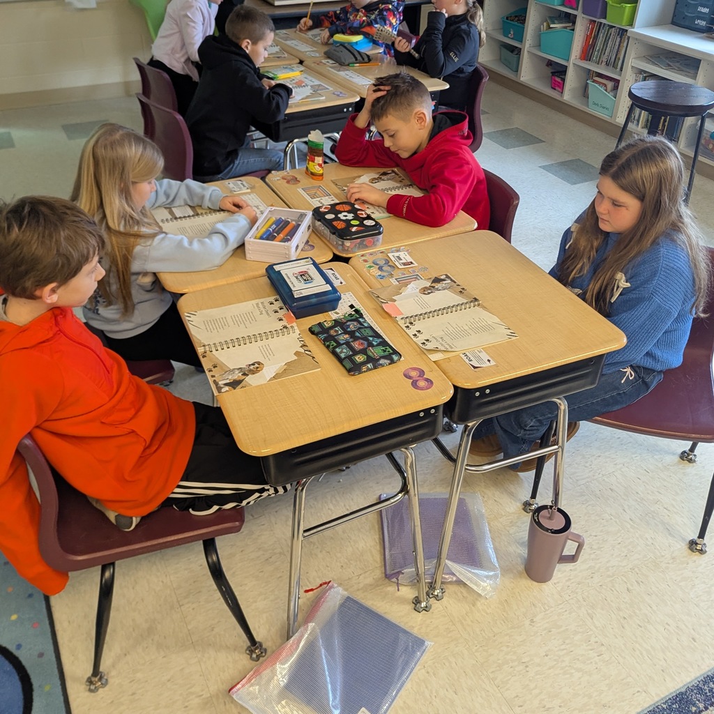 Elementary students sit at grouped desks working on notebooks and sticker activities, surrounded by school supplies and organizers.