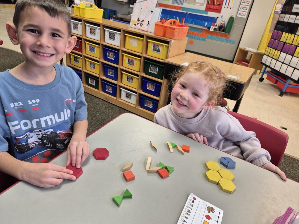 Two smiling children work with various geometric pattern blocks at a table, arranging shapes and colors during a math activity.