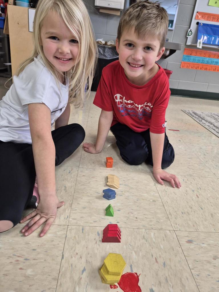 Two children smile while sitting on the floor beside a straight line of stacked pattern blocks sorted by color and shape.