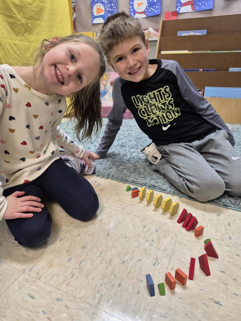 A boy and girl sit on a classroom rug, proudly displaying a curved pattern made of red, yellow, green, and blue blocks.