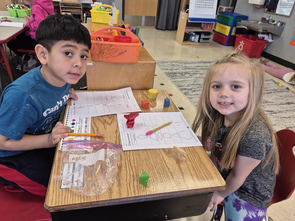 Two young students smile at a classroom table while working on a math activity using pattern blocks and worksheets.