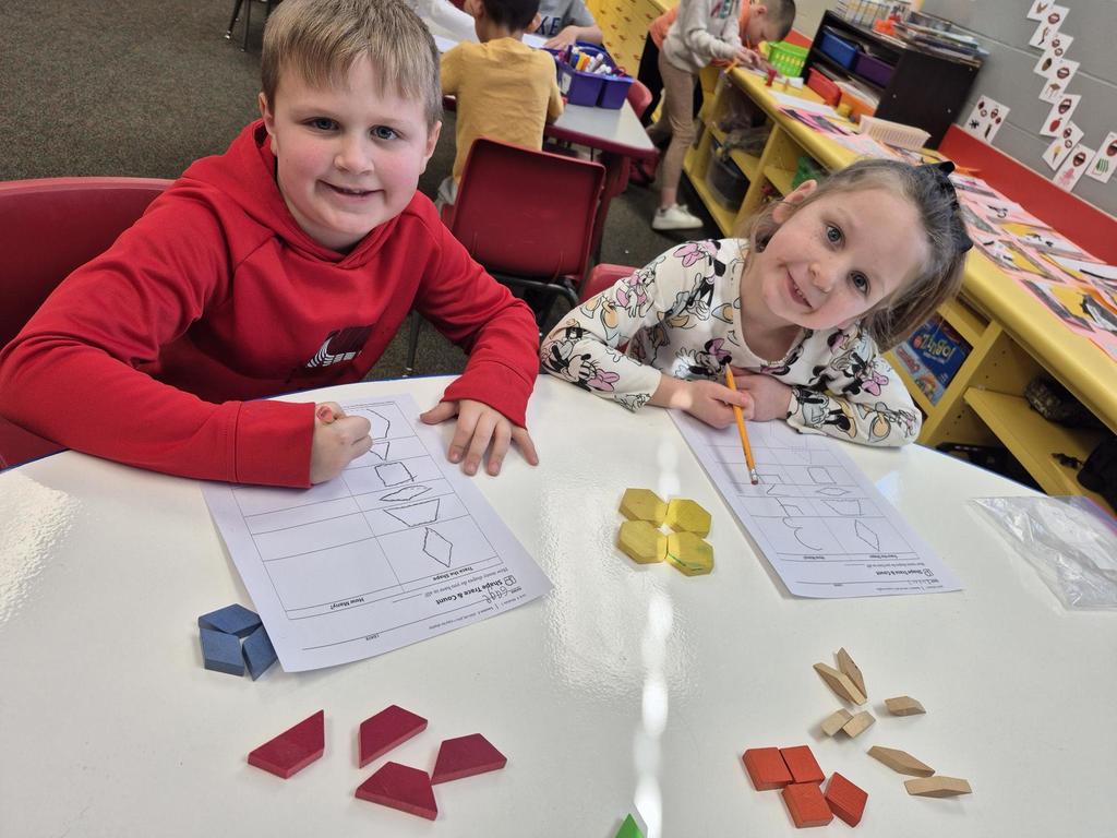 A boy in a red hoodie and a girl in a Minnie Mouse shirt work together at a white table using colorful pattern blocks.