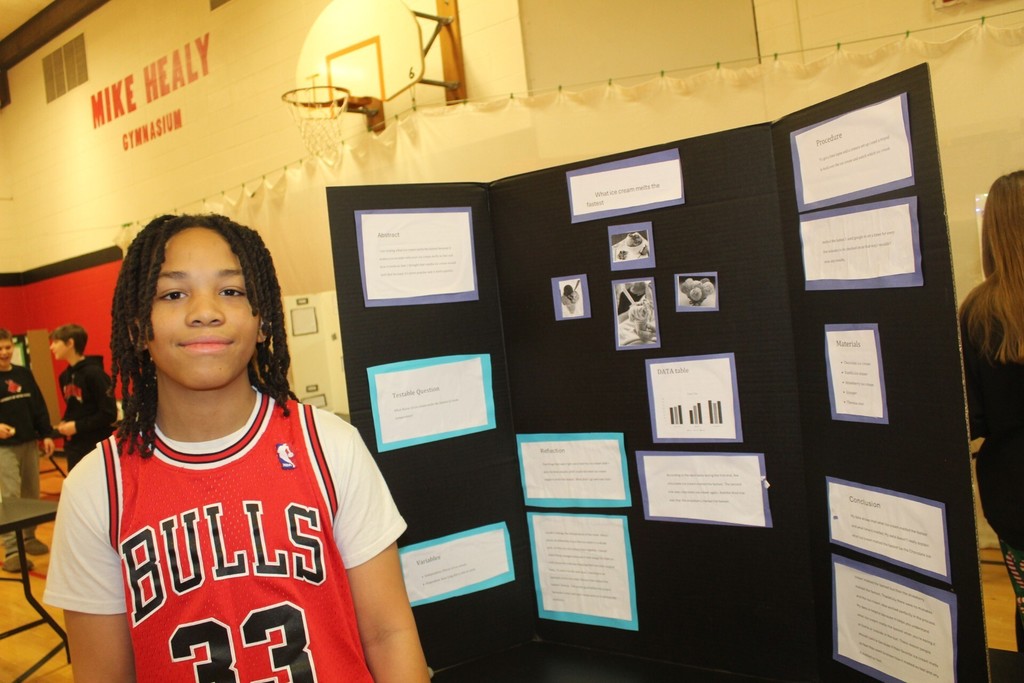 A student stands beside a science fair display board titled “What ice cream melts the fastest,” featuring photos, graphs, and written sections.