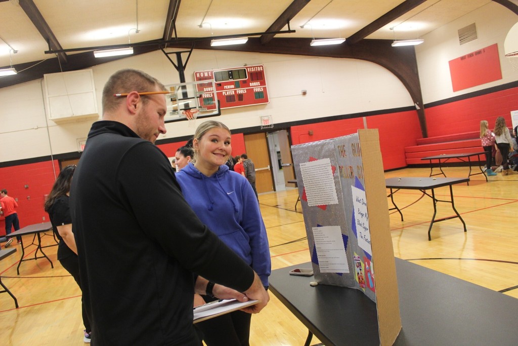 A student in a blue hoodie smiles while presenting a project to an adult holding a clipboard during a school event in the gym.