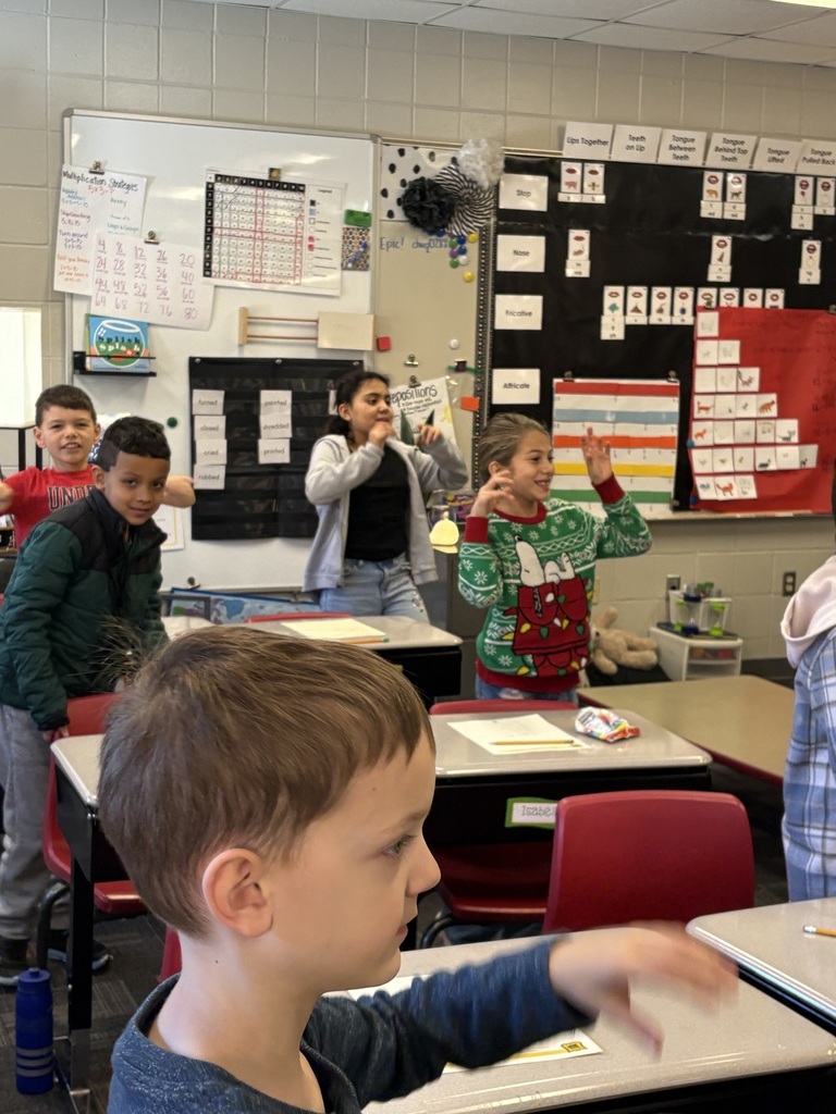 A group of young students stands near their desks participating in an activity together.