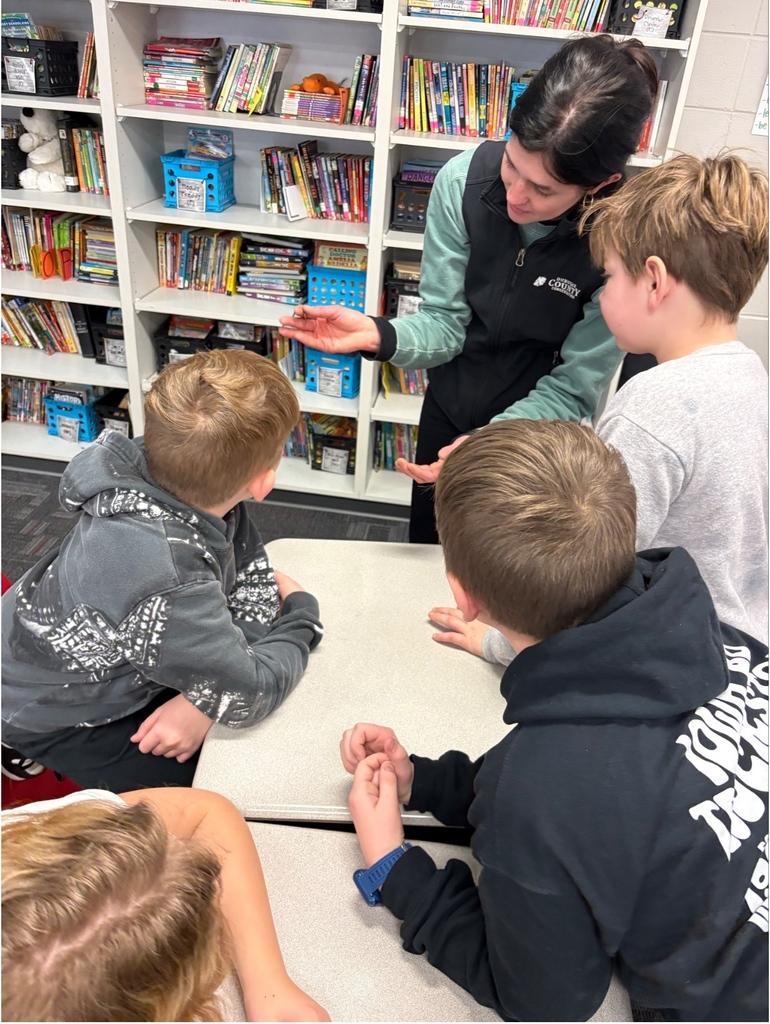 A presenter shows a small snake to a group of students gathered around a desk in front of a bookshelf.