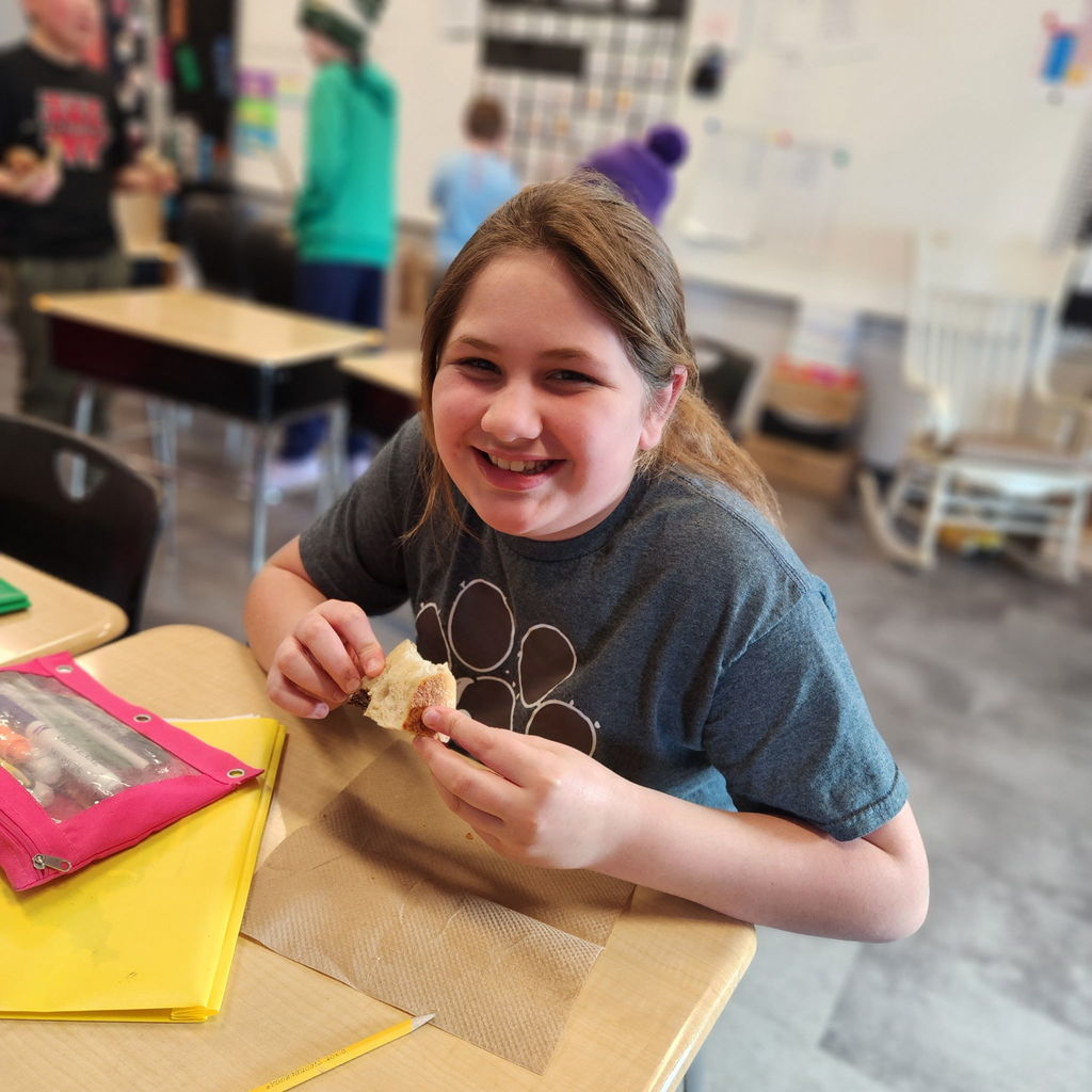 Smiling student sits at a desk enjoying a piece of bread.
