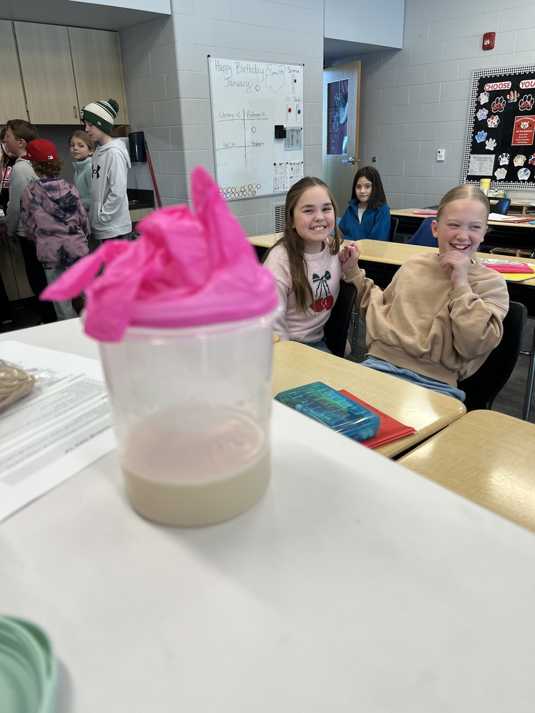 Students sit at desks and smile, with a cup covered by a pink glove in the foreground.