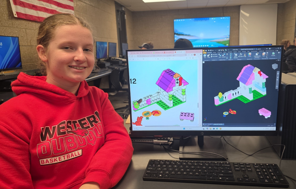 Student poses next to a screen showing a colorful 3D model of a LEGO-style house.