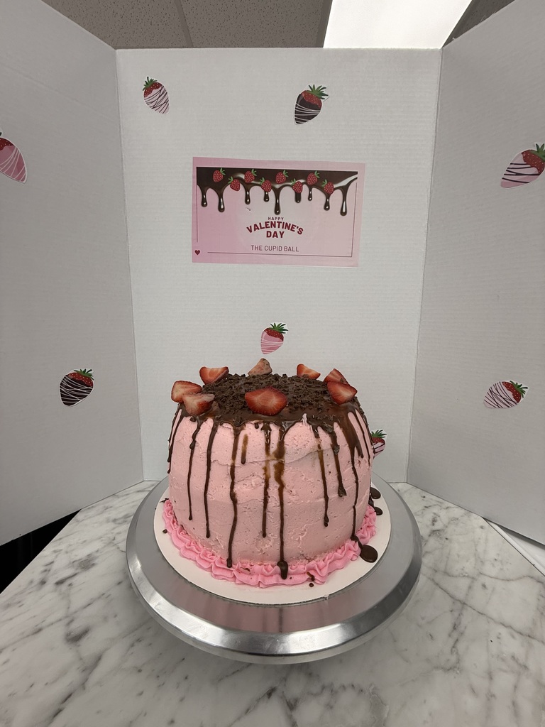Pink cake with chocolate drizzle and strawberries on top, labeled “The Cupid Ball” on a Valentine’s Day-themed display.